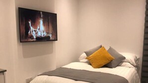 A modern bedroom with a bed, a mounted TV displaying a fireplace, and a neutral color scheme.