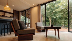 A modern living room with a sofa, armchair, and a small table. There is a kitchen area with a bar counter and stools. Large windows provide a view of the outdoors. - Refugio en la Serva Maya Coba (Cobá)