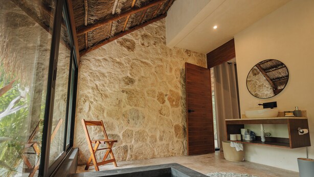 A bathroom with a stone wall, a wooden door, a sink, and a large window. - Refugio en la Serva Maya Coba (Cobá)