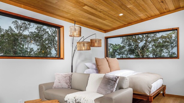 Ein modernes Schlafzimmer mit Holzboden, einem großen Bett mit Holzrahmen, einem beigefarbenen Sofa und zwei großen Fenstern mit Blick auf Bäume.