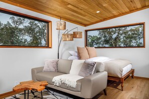 Une chambre moderne avec un plafond en bois, un grand lit doté d’une structure en bois, un canapé beige et deux grandes fenêtres donnant sur la verdure.