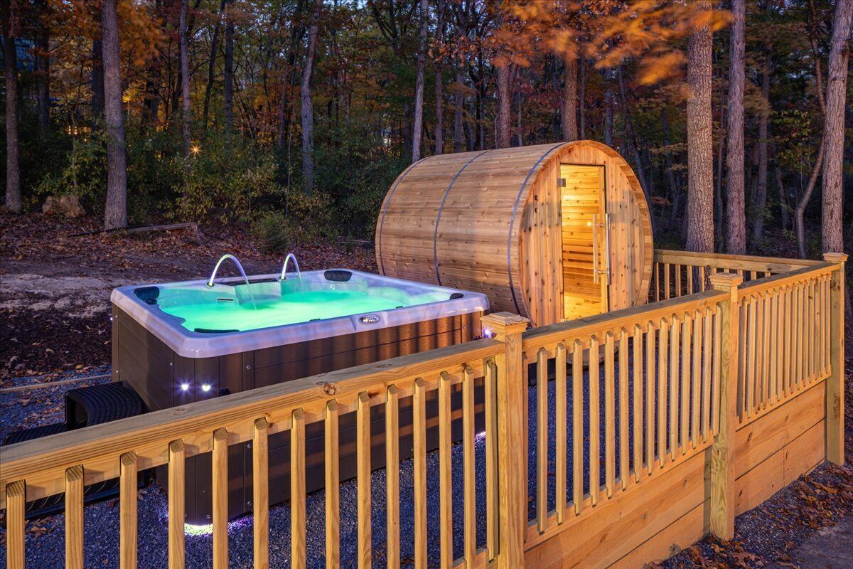 Een hottub met een houten kuipstructuur, omgeven door een houten terras en een omheining, gelegen in een bebost gebied.