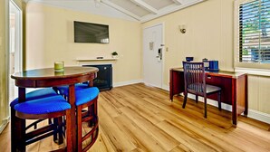 Ein Zimmer mit Holzboden, einem runden Tisch mit blau gepolsterten Stühlen, einem Holztisch mit Stuhl, einem an der Wand montierten Fernseher und einem Fenster mit Jalousien.