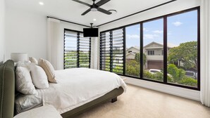 Un dormitorio con una cama grande, un ventilador de techo y vistas a casas vecinas a través de amplios ventanales.