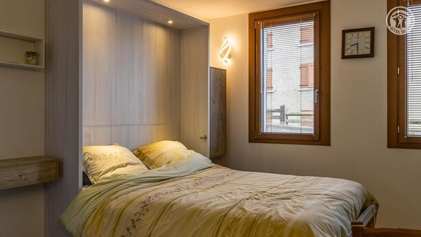 Un dormitorio con una cama, una ventana con persianas, un reloj de pared y una estantería con un tarro.