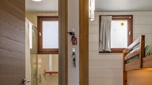 Una habitación pequeña con una litera de madera, un taburete, una ventana con cortinas y un baño con barra para toallas.