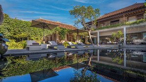 Un espace piscine extérieur luxueux, doté de chaises longues et de parasols, et entouré d’une végétation luxuriante et d’une architecture traditionnelle.