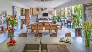 Une cuisine spacieuse dotée d’un îlot central, un coin repas avec des meubles en bois et une vue sur un jardin luxuriant grâce à de grandes fenêtres.