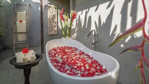 Une salle de bain avec une baignoire sur pieds remplie de pétales de rose, un porte-serviettes avec une serviette et une fleur rouge, une petite table avec des articles de toilette et un panneau décoratif mural.