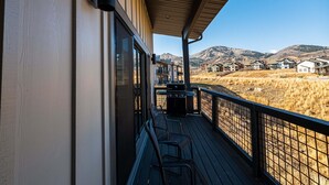 Um deck de madeira com churrasqueira e cadeiras, com vista para uma área residencial com casas e montanhas.