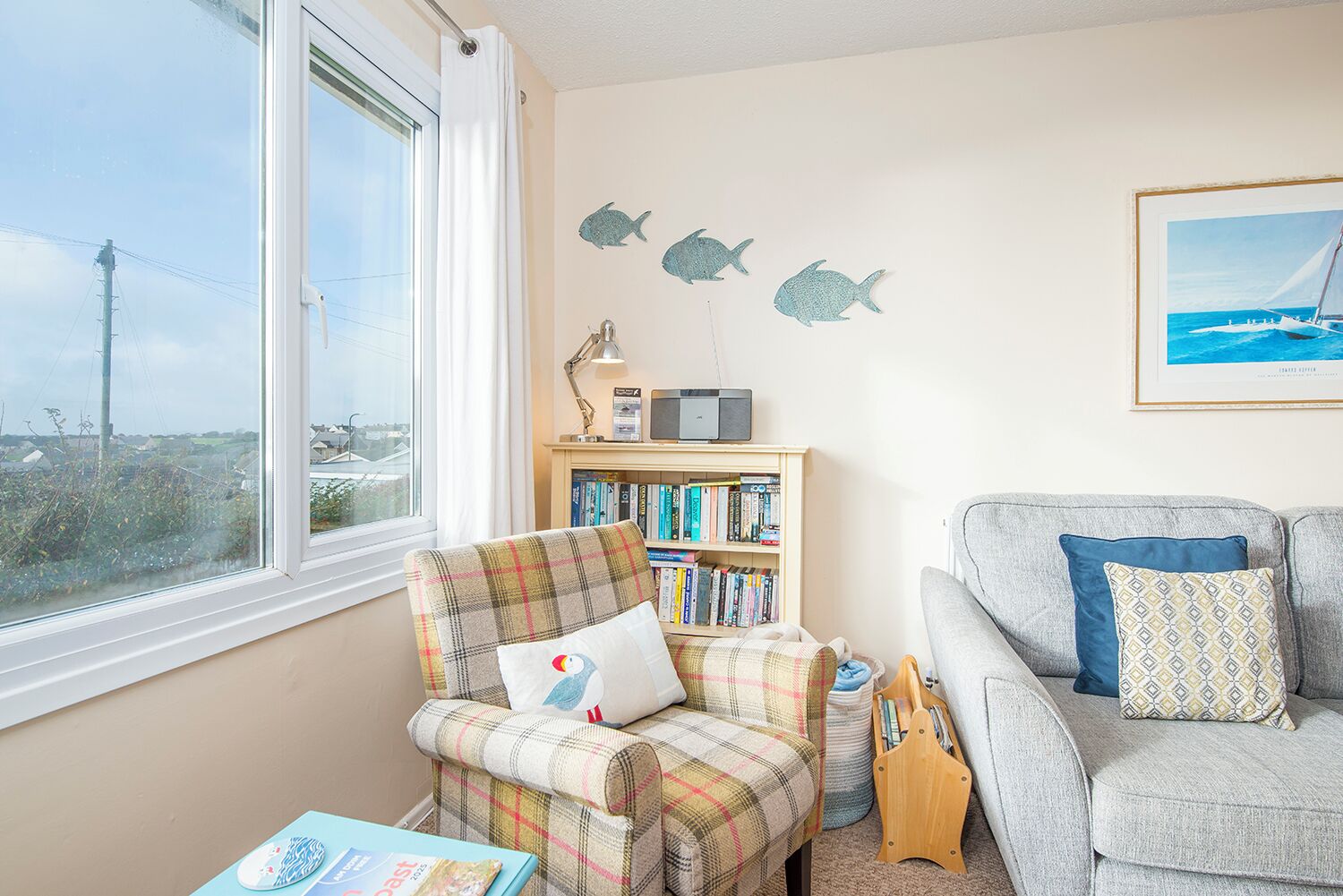 A cozy living room with a large window, a plaid armchair, a bookshelf, a grey sofa, and a framed picture on the wall.