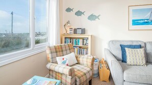 A cozy living room with a large window, a plaid armchair, a bookshelf, a grey sofa, and a framed picture on the wall. - Ty Peggie, Holiday cottage Solva, Sleeps 4, 2 bedrooms,  bathrooms (Haverfordwest)