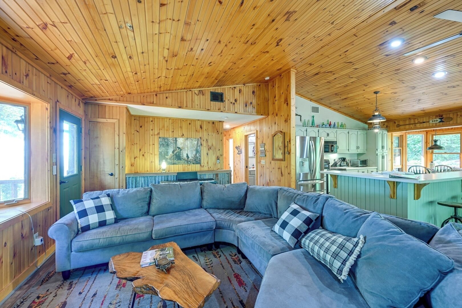 Ein gemütliches Blockhaus-Interieur mit einem großen, grauen L-Sofa, einem Holztisch und einer Küchenzeile mit Kühlschrank und Spüle.