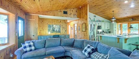 Ein gemütliches Blockhaus-Interieur mit einem großen, grauen L-Sofa, einem Holztisch und einer Küchenzeile mit Kühlschrank und Spüle.