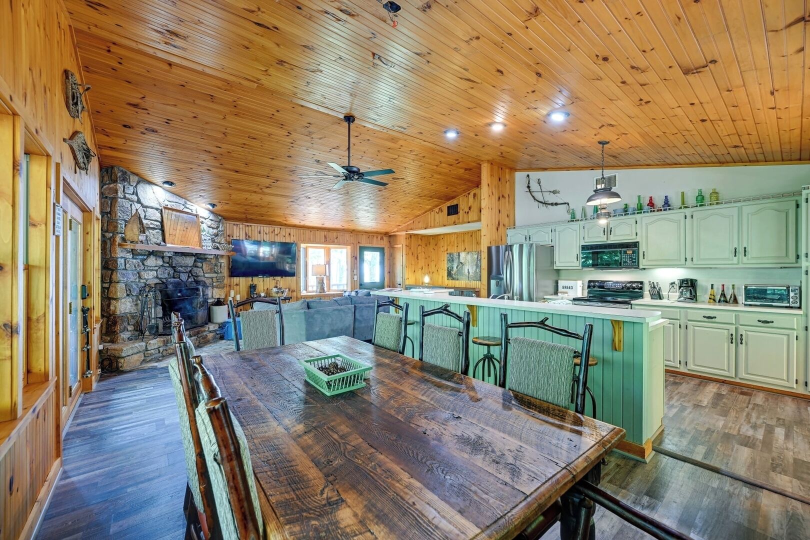 Een knus chaletinterieur met een houten eettafel, een stenen open haard en een keuken met lichtgroene kastjes.