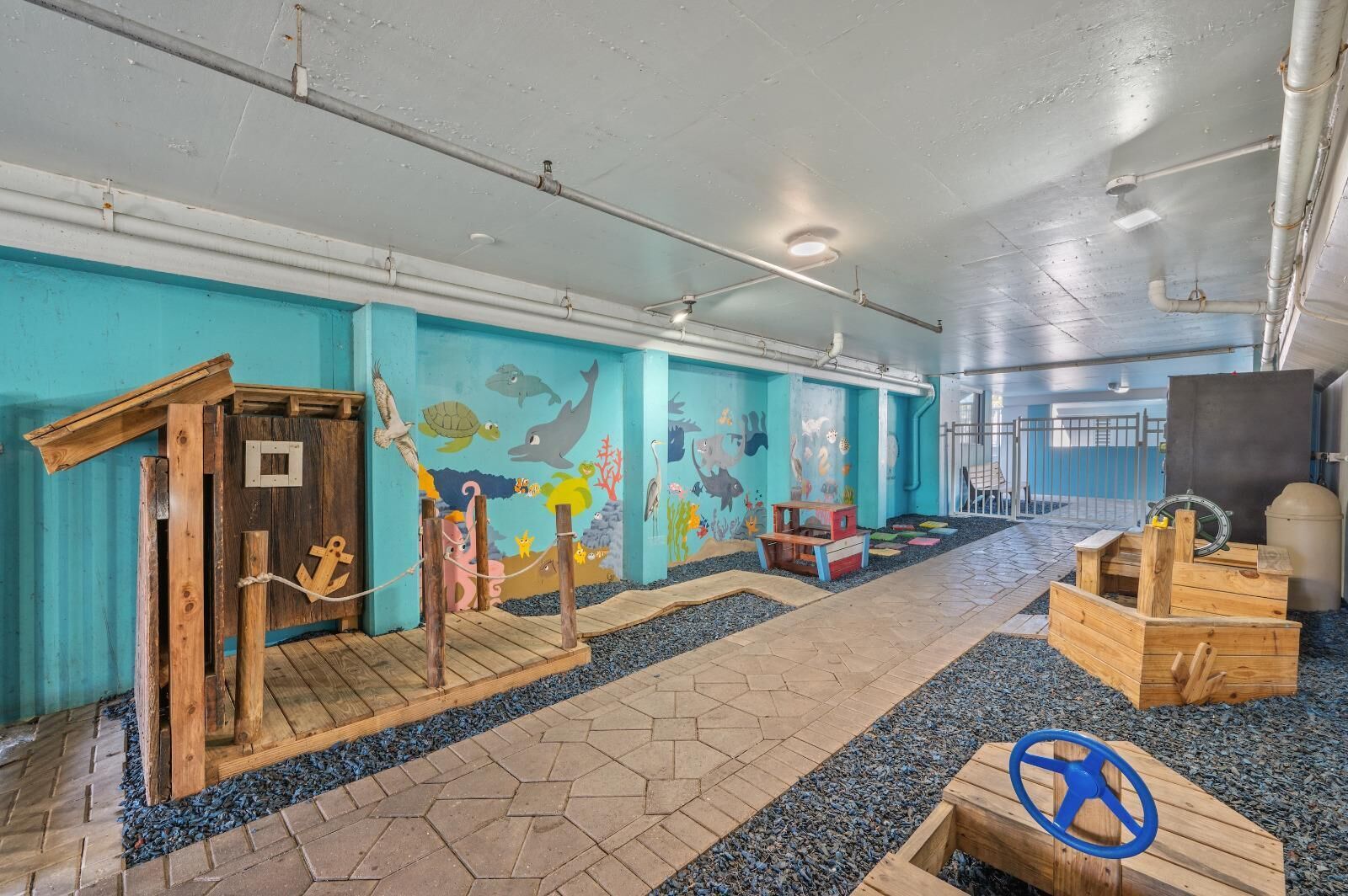 Zona de juegos infantiles con estructuras de madera, un mural de criaturas marinas y suelo de baldosas.