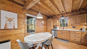 Interior de una cabaña de madera con una mesa redonda para comer, sillas, un reloj y una cocina con armarios y un lavabo.