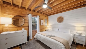 Un dormitorio de cabaña de madera con cama, mesitas de noche, cómoda, ventilador de techo y ventana con cortinas.