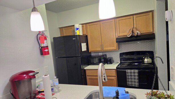 A compact kitchen with wooden cabinets, a black refrigerator, a microwave, and a stove. There is a fire extinguisher mounted on the wall and a small sink with a faucet. - 2-bed apartment, heart of RIO, Gaithersburg! Near 1-270, commuter convenient. (Gaithersburg)