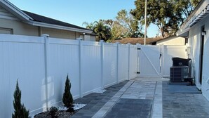 A paved pathway leading to a building with a white fence, small shrubs, and an air conditioning unit. - Perfect terrace to relax, have a coffee or enjoy the Florida weather. (Tampa)