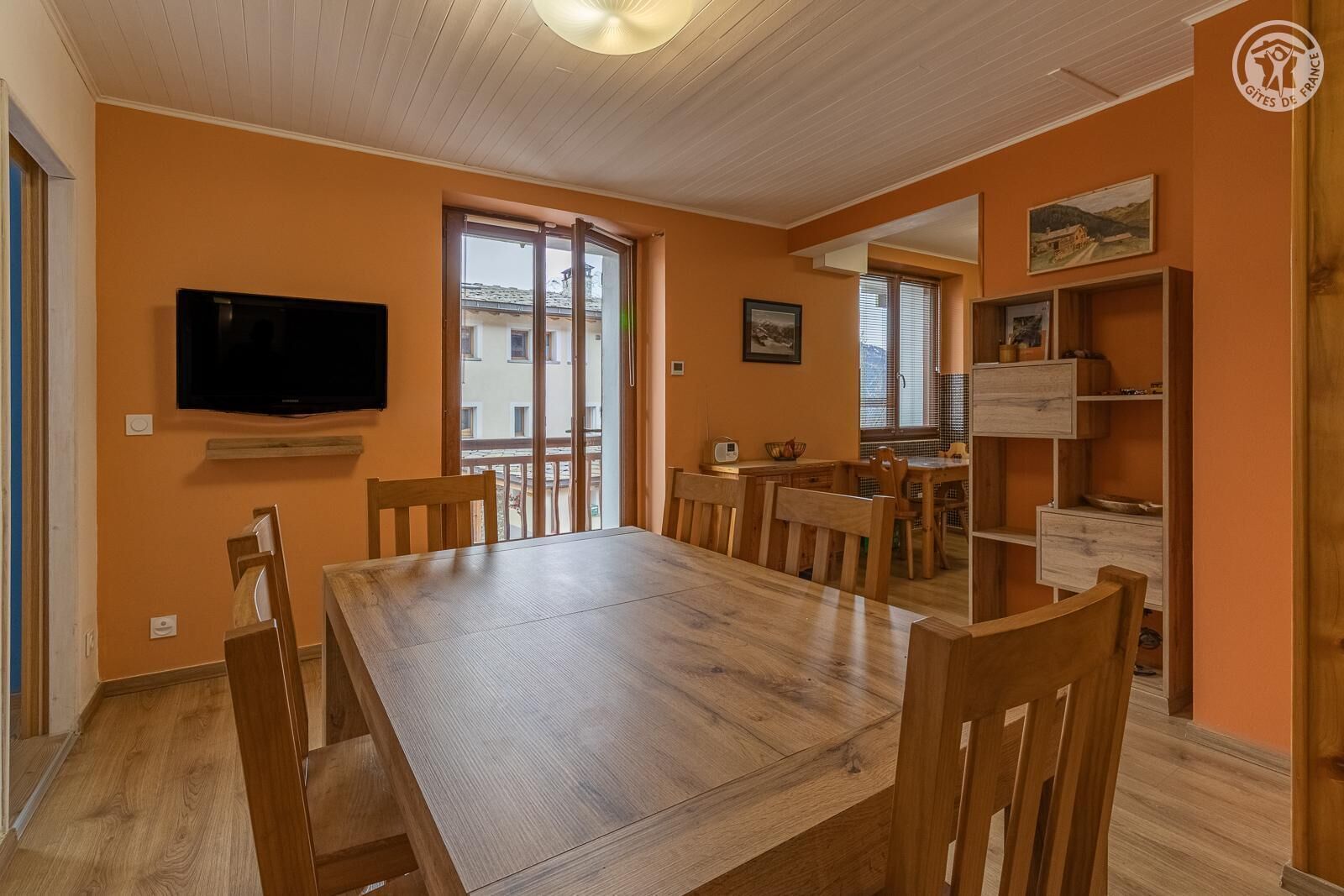 Un coin repas avec une table et des chaises en bois, un téléviseur fixé au mur et une vue sur l’extérieur grâce à une porte coulissante.