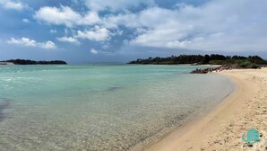 Ein Strand mit klarem türkisblauem Wasser, sandigem Ufer und einer bewaldeten Insel in der Ferne.