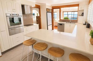 A modern kitchen with a central island, bar stools, stainless steel appliances, and a view of the sea. - Banyandah - home on the water (Marks Point)