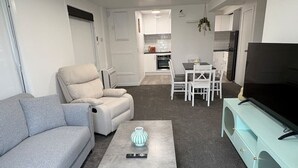 Een moderne woonkamer met een grijze bank, een witte fauteuil, een salontafel met een vaas, en een televisie op een lichtblauwe kast.