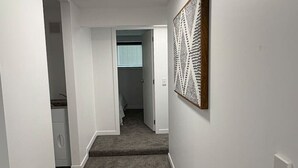 Een hal met een tapijtvloer, witte wanden en een plafondlamp. Aan het einde van de hal bevindt zich een deur die naar een andere kamer leidt.
