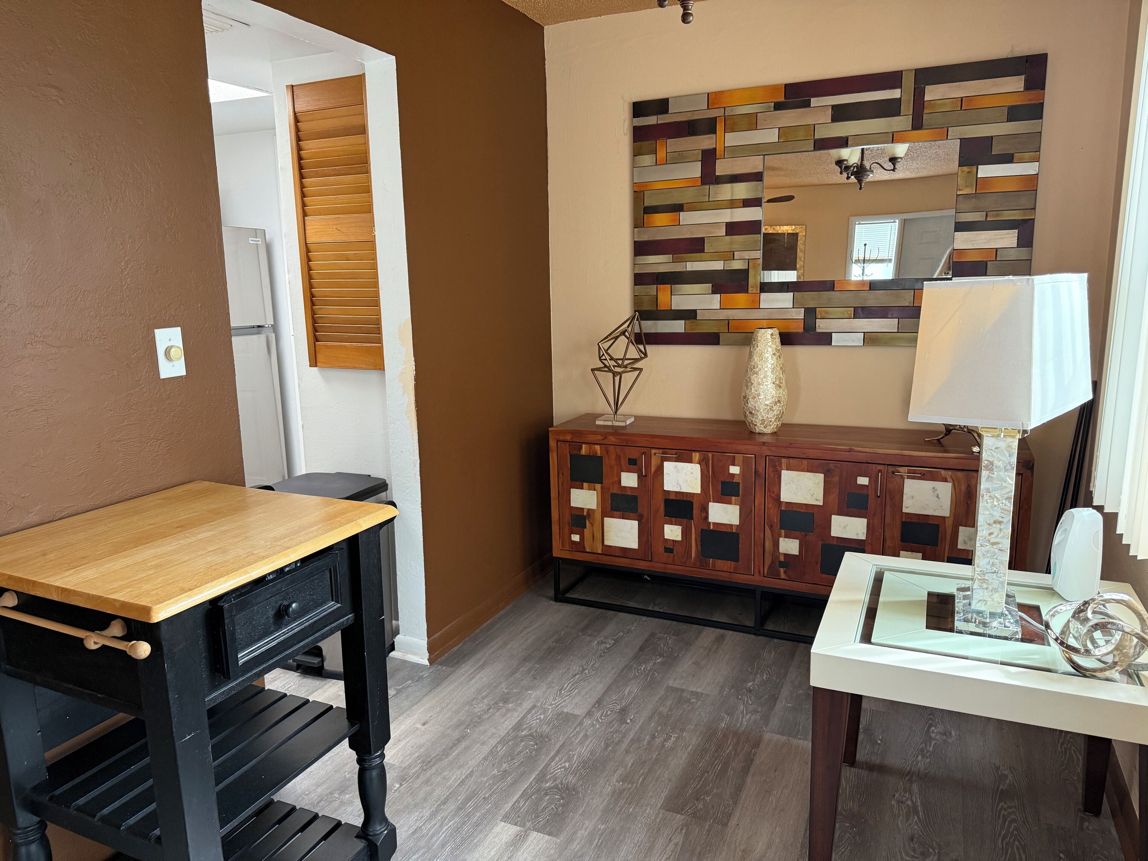 A room with a wooden table, a cabinet with decorative panels, a mirror with a multicolored frame, and a lamp on a side table.