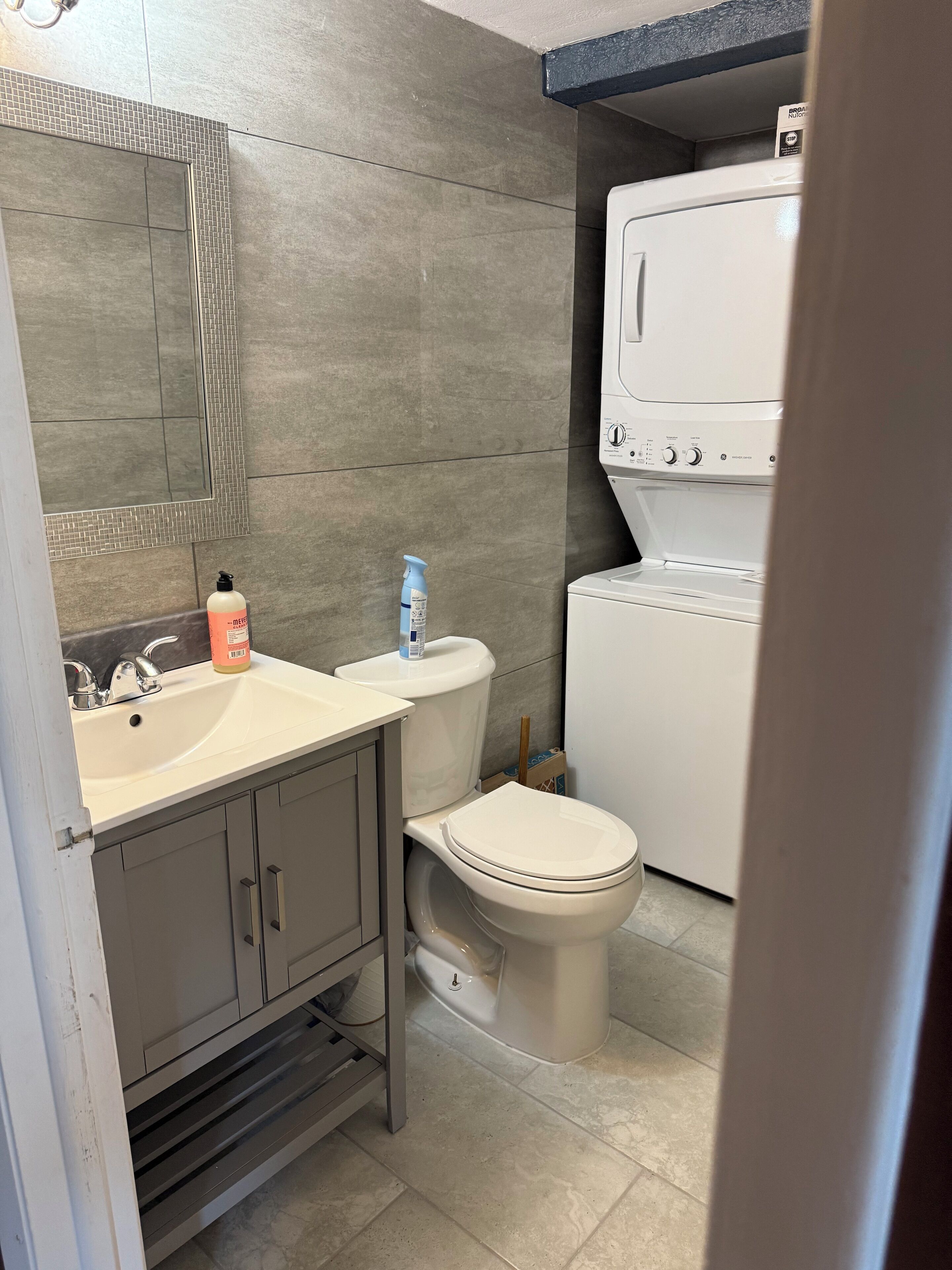 Et badeværelse med en hvid vask, et spejl, et toilet og en vaskemaskine.