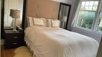 A neatly made bed with a headboard, a bedside table with a lamp, and a view of greenery outside.