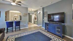 Een moderne woonkamer met een grote flatscreen televisie, een plafondventilator en een eethoek met twee blauwe stoelen.