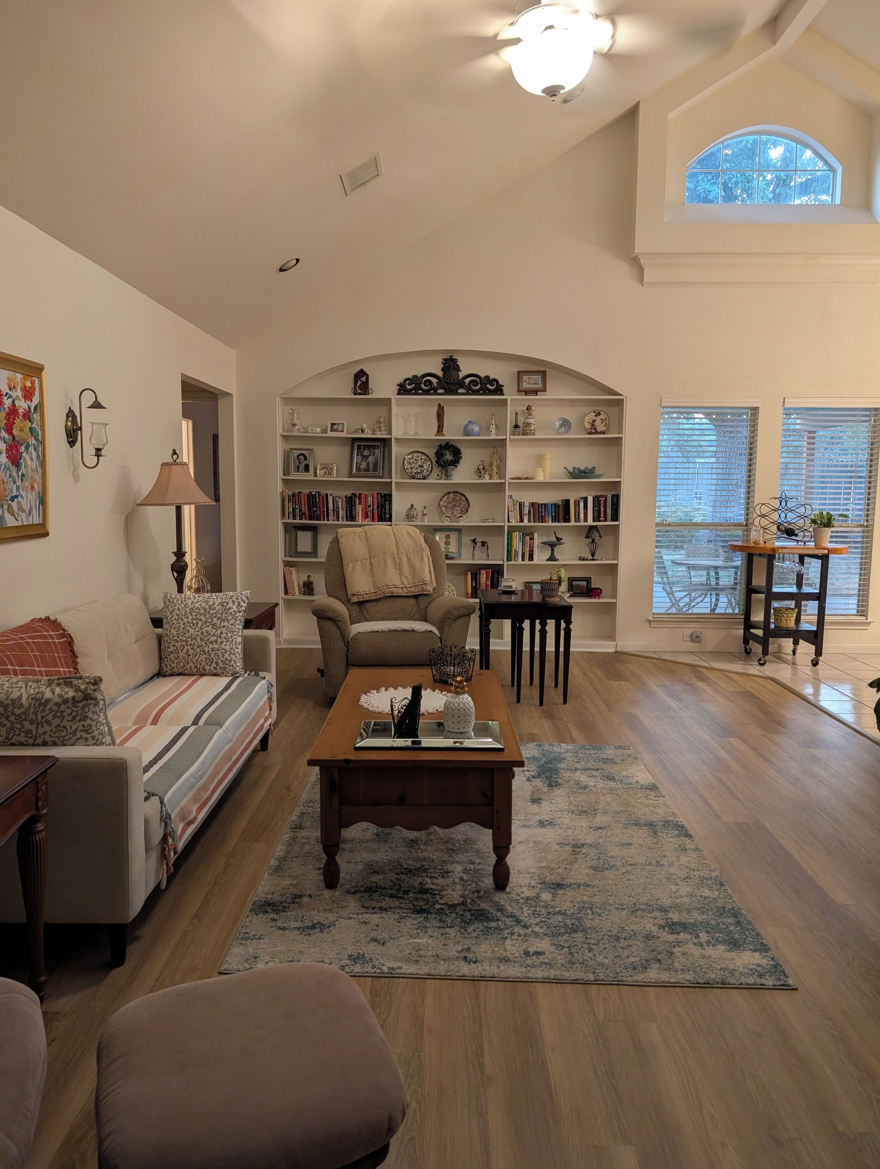 A living room with a sofa, armchair, coffee table, bookshelf, and a view of the outdoors through a window.