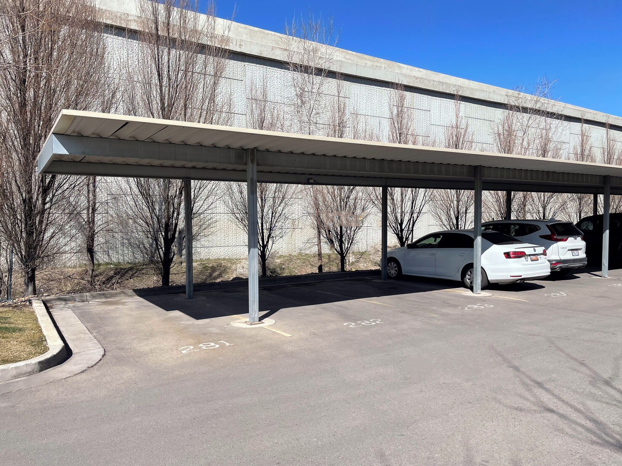 A covered parking area with two cars parked, a concrete wall in the background, and a clear sky.