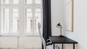 A room with a wooden floor, a white radiator, a black desk with a lamp, a grey chair, and a window with white shutters. - Blueground | Wieden, view, nr Belvedere (Vienna)