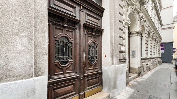 A building with two large wooden doors with intricate metalwork, a stone facade, and a narrow sidewalk. - Blueground | Wieden, view, nr Belvedere (Vienna)