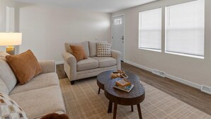 A living room with a sofa, a round coffee table, and a lamp. There are two windows with blinds and a door.