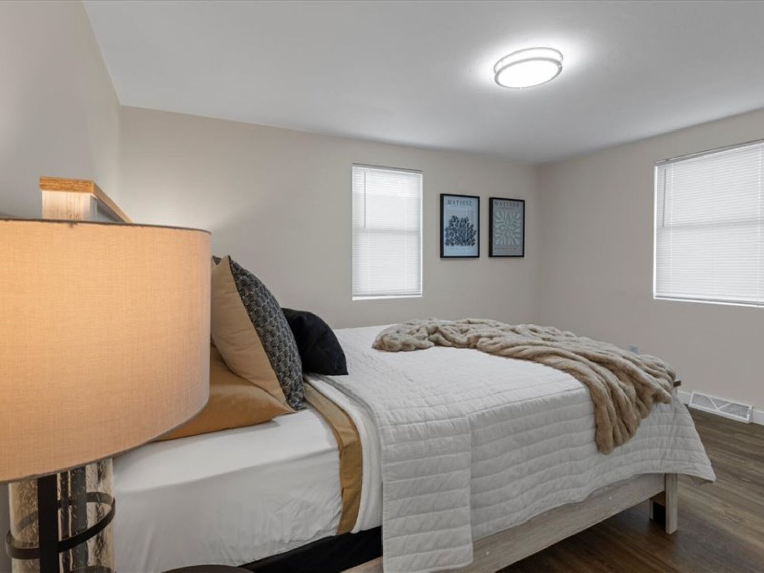 A bedroom with a bed, a bedside lamp, two framed pictures on the wall, and a window with blinds.