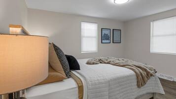 A bedroom with a bed, a bedside lamp, two framed pictures on the wall, and a window with blinds.