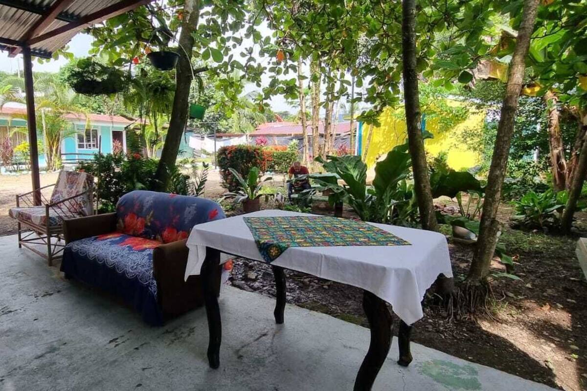 An outdoor seating area with a table and a sofa, surrounded by lush greenery and a building in the background.