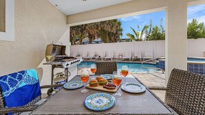 Un área de comedor junto a la piscina con una parrilla, sillones de descanso y una mesa dispuesta con platos, vasos y comida.