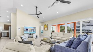 Amplia sala de estar con sofás beige y azules, una mesa de centro y un televisor. Hay ventiladores de techo, una cocina al fondo y un balcón con vistas.