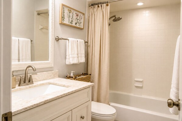 A bathroom with a sink, mirror, toilet, shower, and a framed botanical print on the wall. - Your home away from home II (New Port Richey)