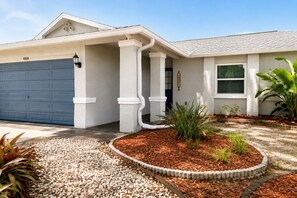 A residential house with a garage, a covered porch, and a landscaped front yard with mulch and plants. - Your home away from home II (New Port Richey)