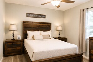A bedroom with a wooden headboard, two bedside tables with lamps, a bed with white bedding, a ceiling fan, and a wall-mounted sign. - Your home away from home II (New Port Richey)