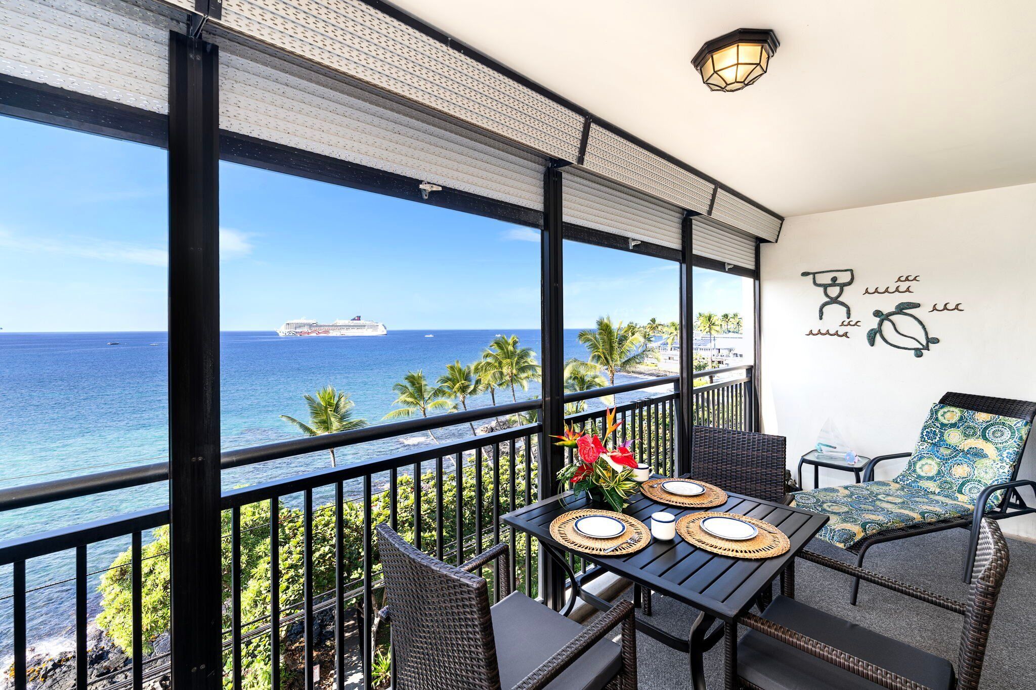 A balcony with a view of the ocean and a cruise ship, furnished with wicker chairs and a table set for two.