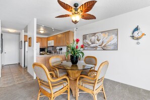 Ein Essbereich mit einem Tisch mit Glasplatte, umringt von Korbstühlen, einem Deckenventilator, einer Kübelpflanze mit Blumen und einer Wanduhr.