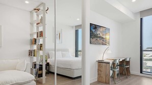 Quarto moderno com cama, guarda-roupa branco, escrivaninha com cadeiras e uma janela grande com vista.