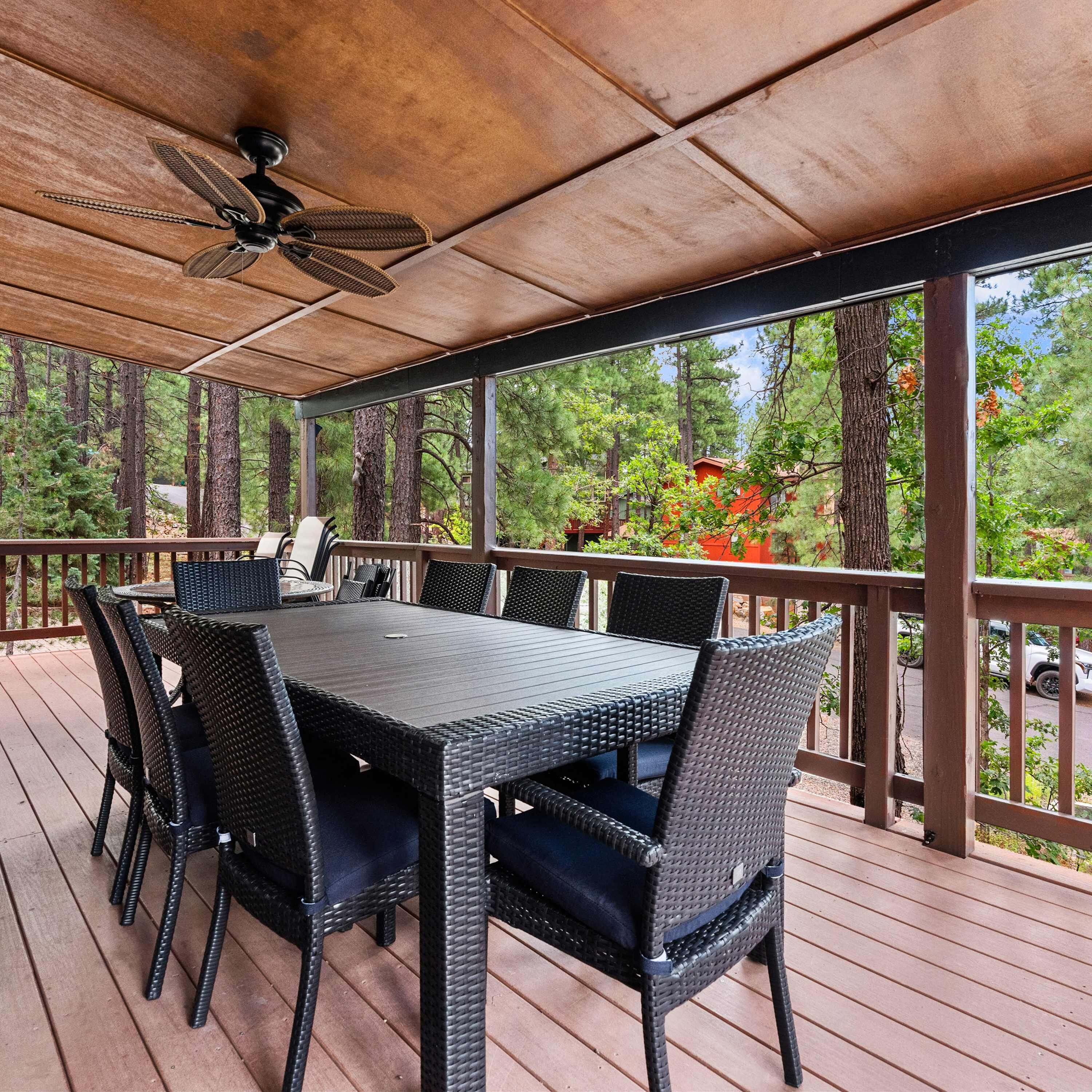 Eine überdachte Terrasse im Freien mit Tisch und Stühlen, einem Deckenventilator und Blick auf Bäume und ein Gebäude.
